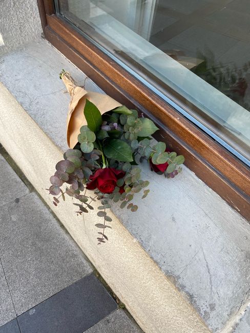 A small bouquet of fresh red roses and eucalyptus sprigs, wrapped in beige paper, rests against a windowsill on a concrete ledge outside a building. The roses have vibrant, velvety petals, and the euc