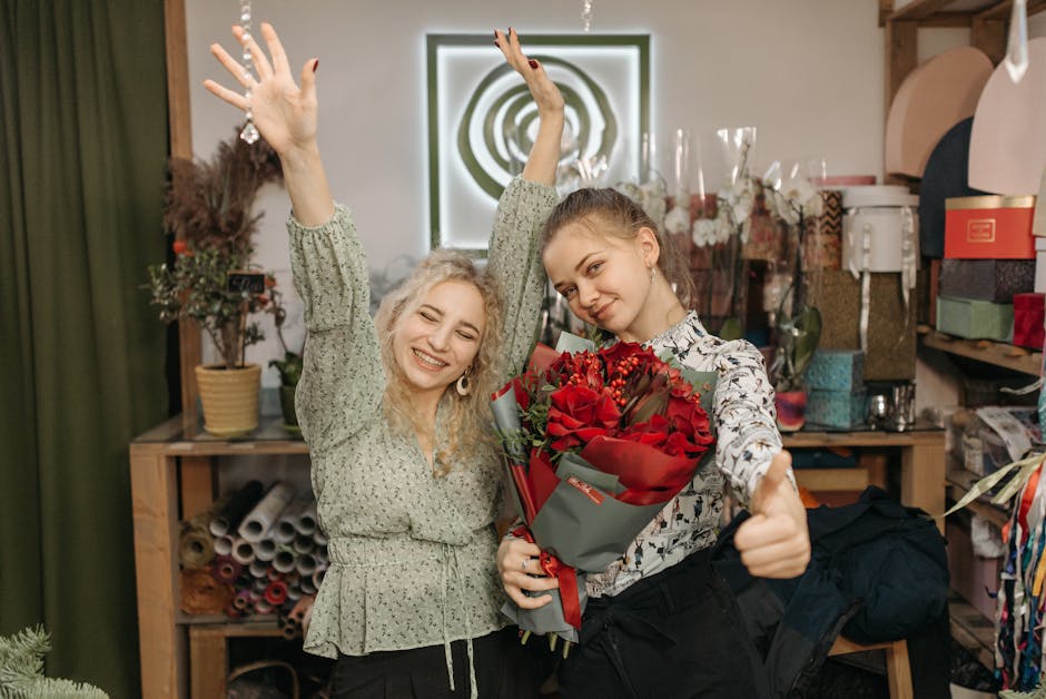 Two young women inside a flower shop, with one holding a bouquet of vivid red roses wrapped in grey paper tied with a red ribbon, both smiling brightly. The woman on the left has curly blonde hair and