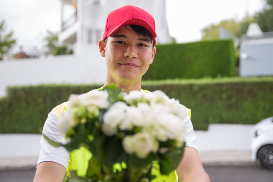 A young delivery person wearing a red cap and a high-visibility vest is holding a bouquet of fresh white flowers, which includes roses and possibly peonies or ranunculus, wrapped in clear tissue and p