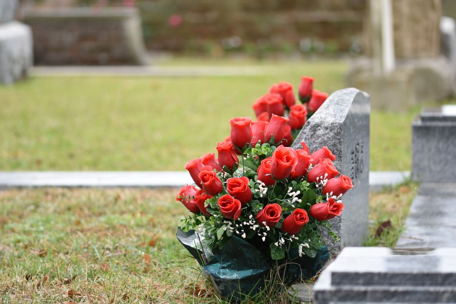 A floral tribute featuring a bouquet of vibrant red roses arranged with small white filler flowers and green foliage, wrapped in dark green paper. The bouquet is placed leaning against a gray stone gr