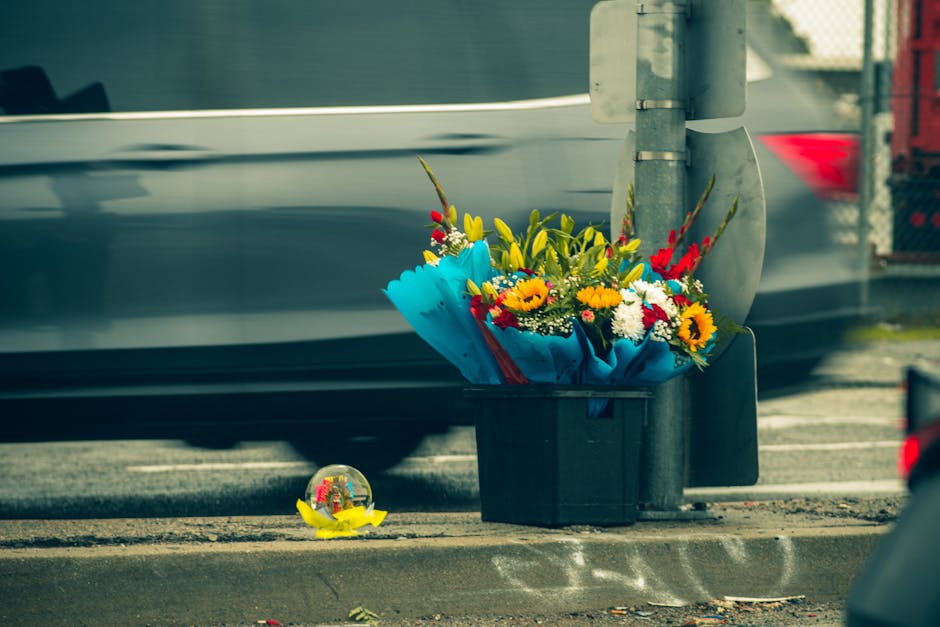 A roadside floral tribute featuring a vibrant bouquet of fresh flowers, including yellow sunflowers, red and white blooms, and green foliage, arranged in a wrapping of bright blue paper and placed in 
