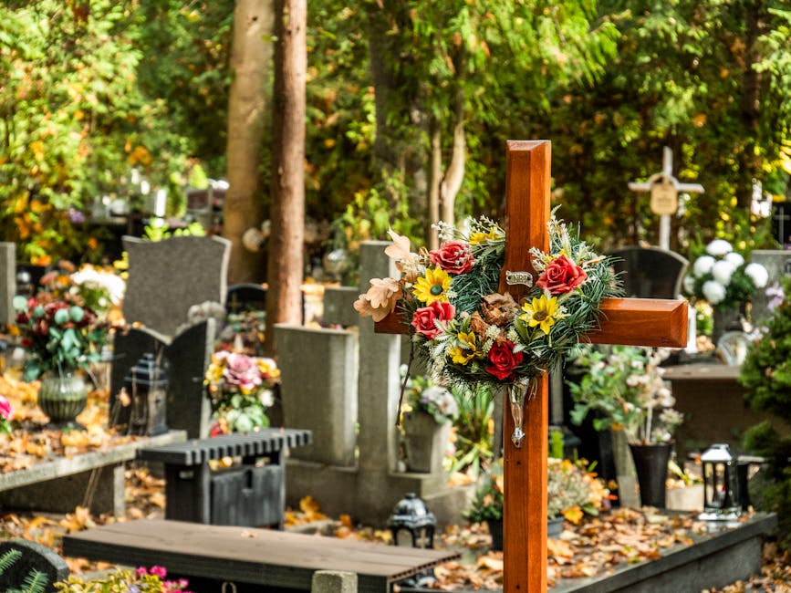 A wooden cross decorated with a floral arrangement consisting of red roses, yellow daisies, pink lilies, and green foliage, placed among graves in a cemetery. The background features headstones and me