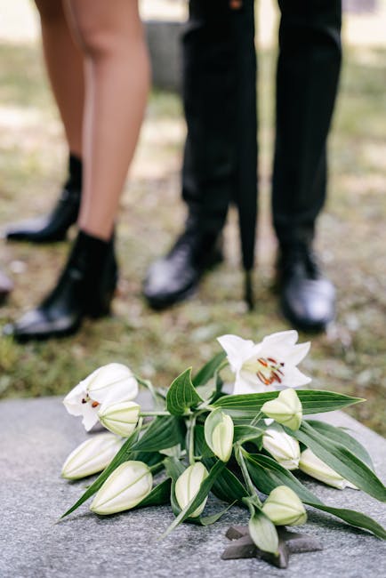 A bouquet of fresh white lilies with large, open petals and prominent orange stamens, along with several closed lily buds and green leaves, is placed on the ground in front of two people dressed in fo