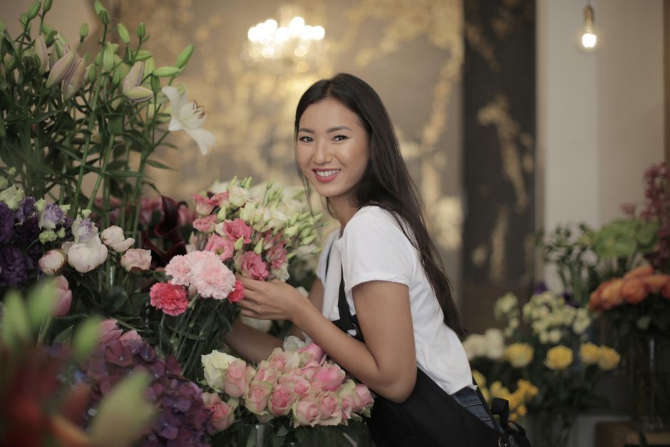 A young woman with long dark hair and a friendly smile is holding a bouquet of pink and white flowers, including roses and carnations, inside a floral shop. She is wearing a white T-shirt and an apron